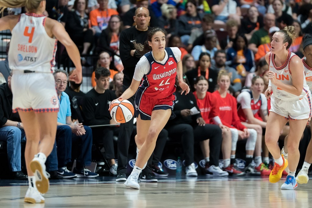 Washington Mystics guard Sonia Citron dribbles the ball with her right hand as a Connecticut Sun defender chases her. Citron's eyes are up, and Mystics head coach Sydney Johnson is visible in the background.