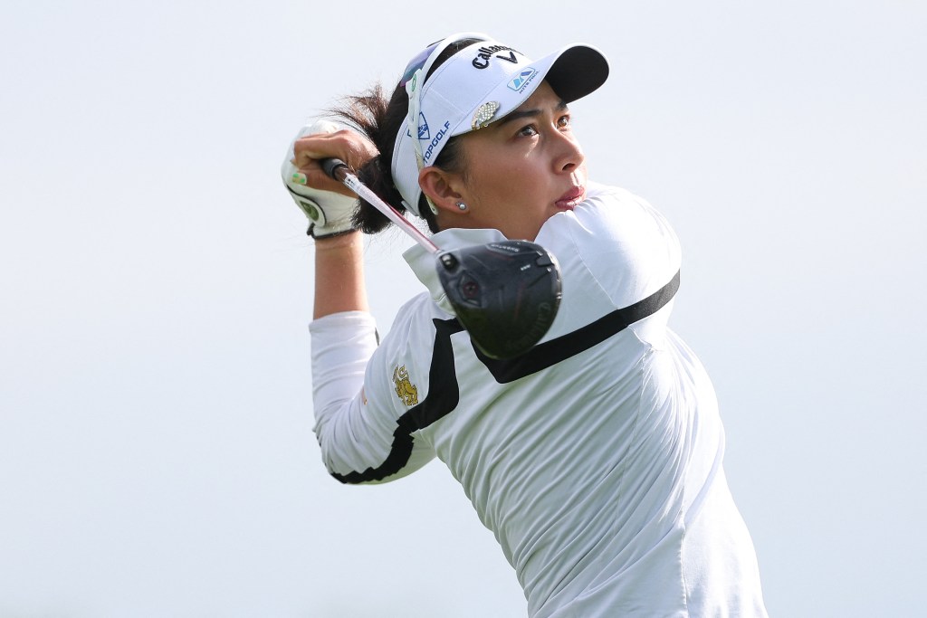 Jeeno Thitikul hits a golf tee shot and looks after the ball. Her hands and the shaft of the club are behind her head in a follow-through position.