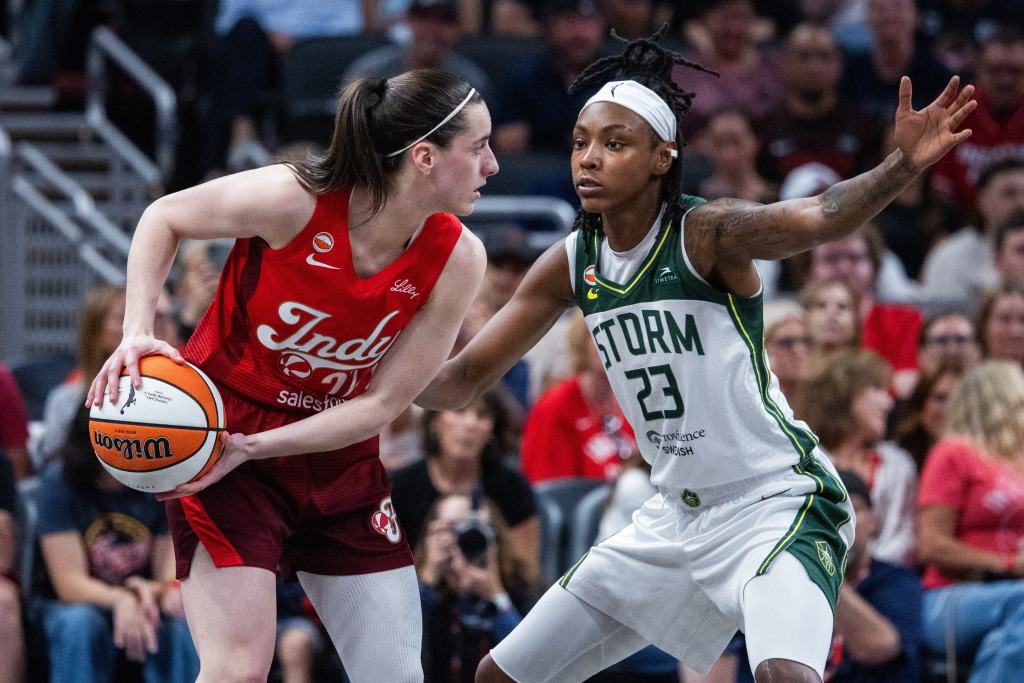 A player in a red uniform holds on to the basketball while a player in a green and white uniform attempt to stop her. 