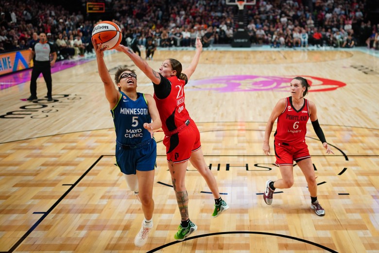 Olivia Miles in her blue Lynx uniform jumps towards a basketball hoop against two Portland defenders in red backdropped by a basketball court