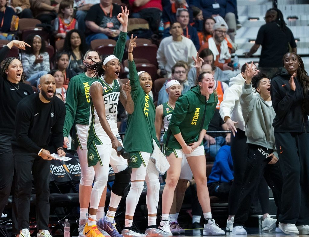 Jordan Horston expresses shock and joy as teammates around her scream and hold up three fingers to celebrate a teammate on the floor scoring a 3-pointer.