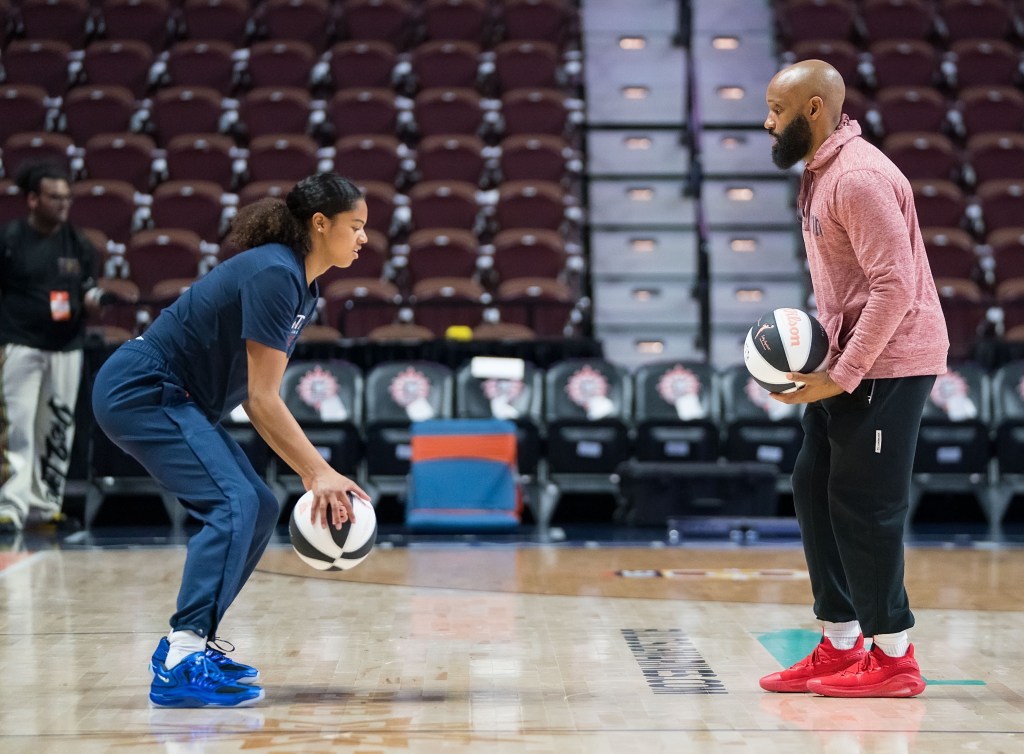 Washington Mystics guard Cassandre Prosper and player development lead Clinton Crouch are shown in profile, facing one another and each holding a basketball. Prosper bends her knees and dribbles the ball as Crouch observes.