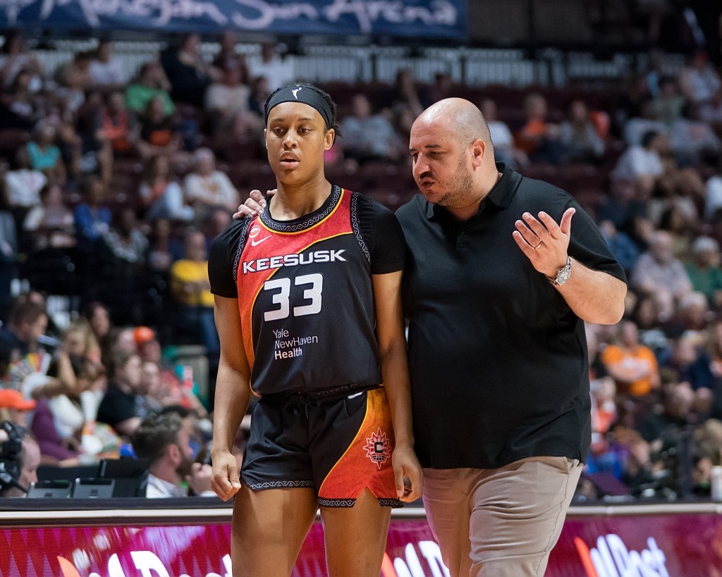 Connectcut Sun head coach Rachid Meziane with his arm around guard Nell Angloma while coaching her on the sideline. Meziane is wearing a black shirt with tan pants and Angloma is wearing a black No. 33 Sun jersey with orange and red accents.