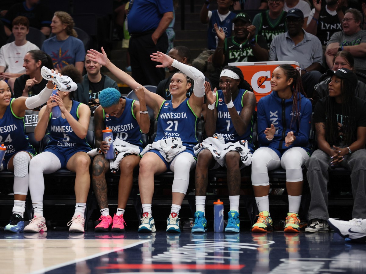 Kayla McBride sits on the bench in the middle of her Lynx teammates in blue uniforms. She's waving her arms in the air and has a big smile on her face.