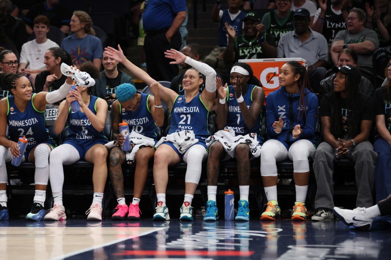 Kayla McBride sits on the bench in the middle of her Lynx teammates in blue uniforms. She's waving her arms in the air and has a big smile on her face.