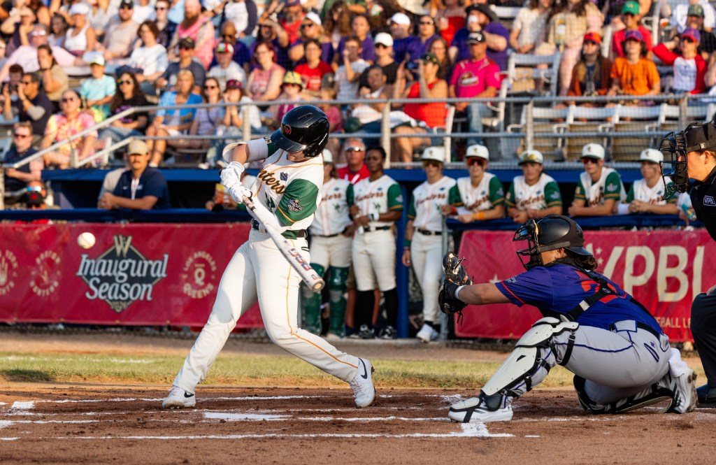 On a sunny day at Robin Roberts Stadium, a left-handed-hitting Boston Hunters batter swings a white bat. She wears a white uniform with orange trim and green sleeves, a black batting helmet, and white batting gloves and spikes swings. The ball, heading left to right, is in frame on the left. Firebells catcher Alexia Jorge, in a purple jersey with orange trim and grey pants with purple trim, holds her black glove out to receive the pitch on the right of the image. Behind them is the Hunters dugout and the stands. The front of the dugout is draped with red WPBL banners, and seven Hunters players stand in the dugout watching the action.