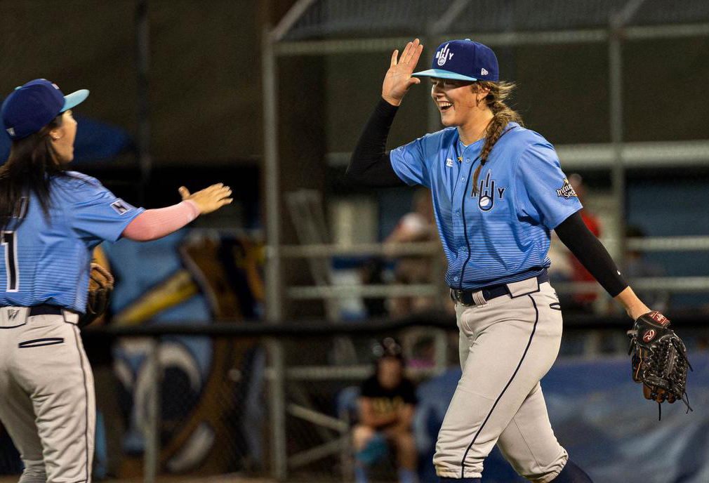 With a big smile on her face, Heights pitcher Jaida Lee, on the right of the photo, holds her right hand up for a high-five with teammate Keira Izumi, who is facing away from the camera on the left. They both wear navy caps, sky blue jerseys, and grey pants.