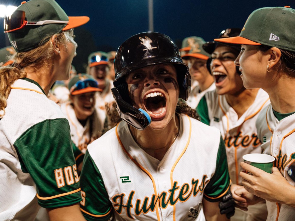 Hunters outfielder Lexi Hastings screams into the camera while wearing a black batting helmet with a C-flap on the left, streaks of eye black down her cheeks, and a white jersey with green sleeves and orange trim. Her teammates celebrate behind her.