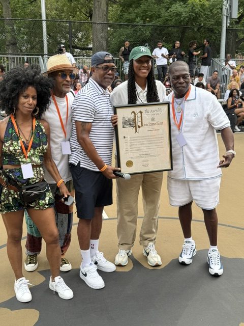 A smiling woman holding a large proclamation is surrounded by three men and a woman. One of the men is holding the microphone backwards and wearing his hat backwards.