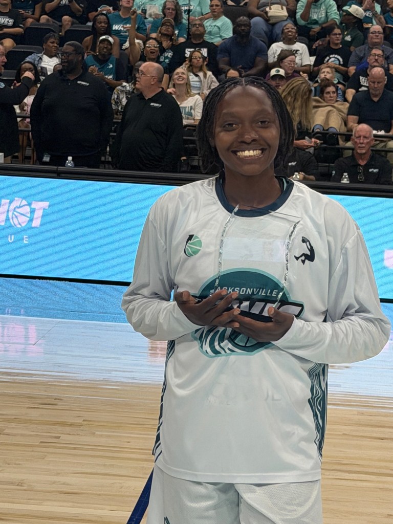 Holding a clear trophy, Ariel Hearn is wearing a white warmup shirt and white shorts as a crowd sits in the background.