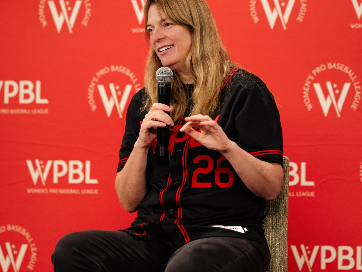 WPBL commissioner Justine Siegal sits on a tall chair in front of a red backdrop with the WPBL logo repeating in white. She holds a microphone in her right hand and gestures with her right. She is wearing black pants and a black WPBL jersey with red trim and the number 26 on the lower left (from her perspective).