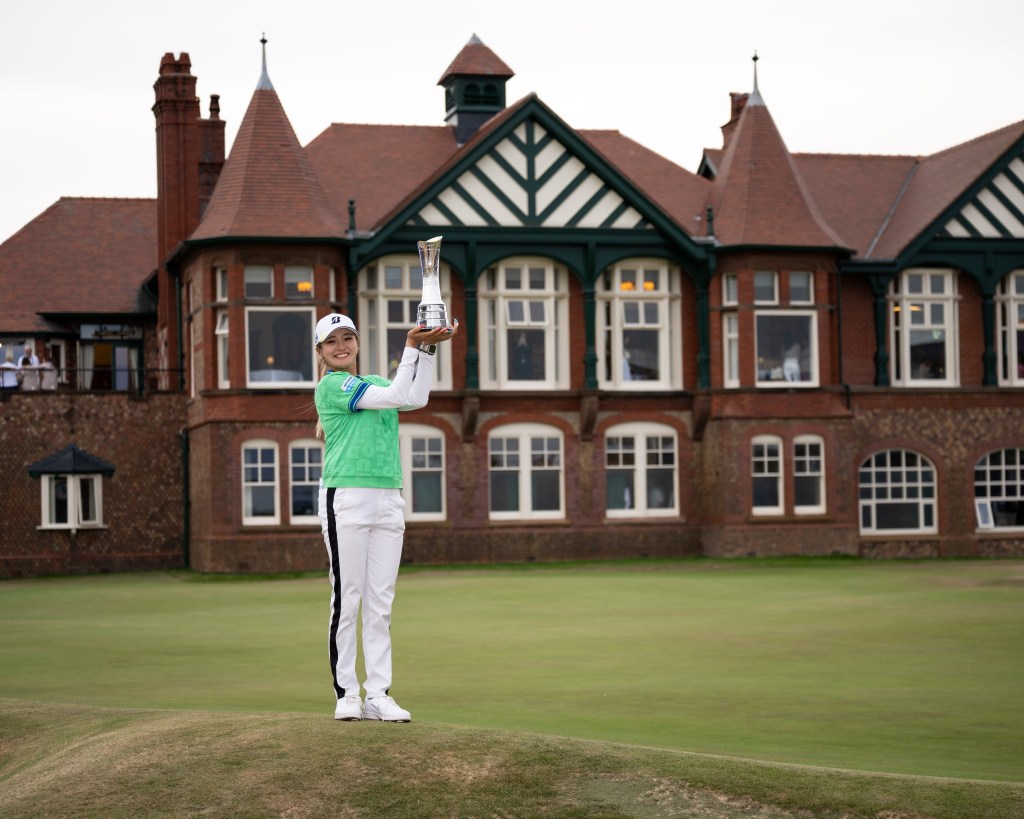 Shiho Kuwaki stands on the edge of the 18th green holding up the AIG Women's Open trophy to her left. The iconic Royal Lytham & St Annes clubhouse is the backdrop.