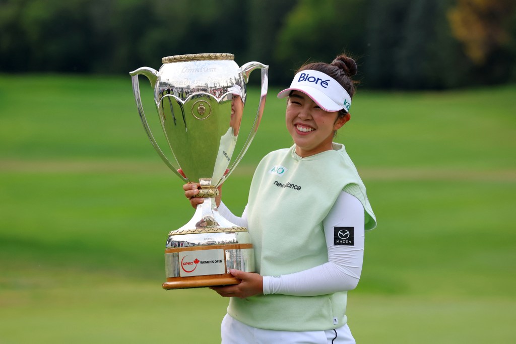Miyu Yamashita holds up the large, silver CPKC Women's Open trophy to her right. Her right hand grabs the trophy and her left hand supports the base. She smiles widely.