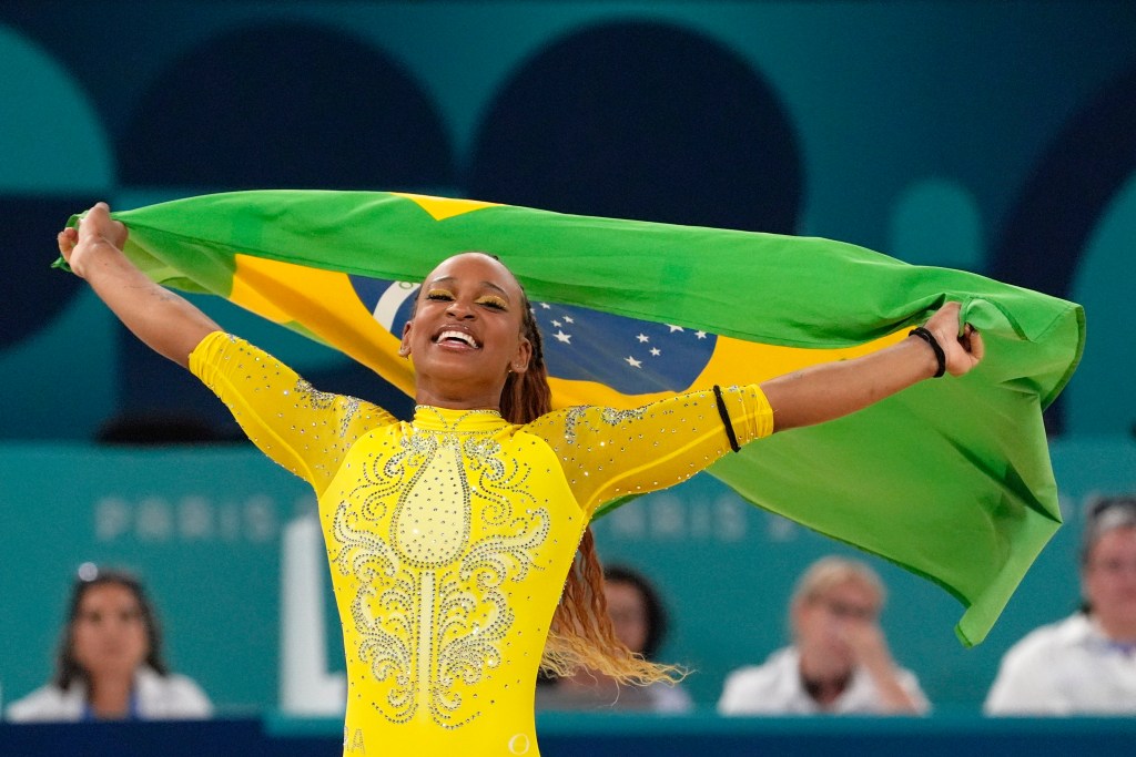 Rebeca Andrade wears a gold leotard and holds Brazil's flag behind her with arms outstretched and smiling in celebration