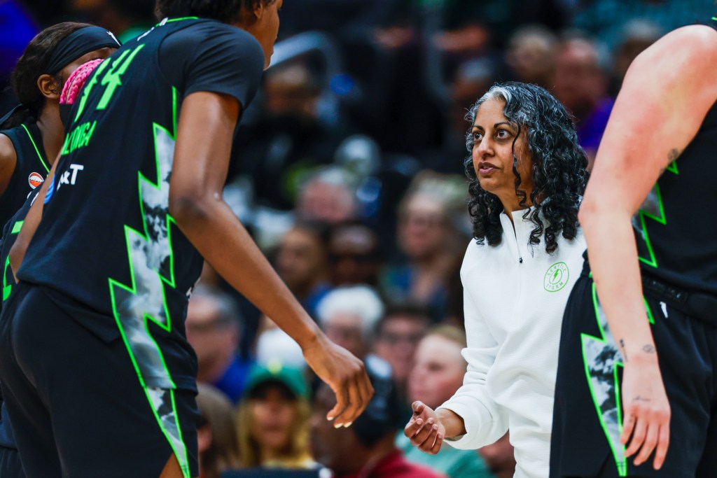 A woman in a white quarter zip coaches players on the court in green and black
