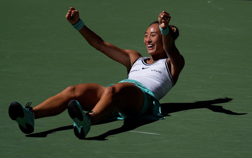 Qinwen Zheng smiles and throws her arms up in the air in celebration while sitting on a tennis court.