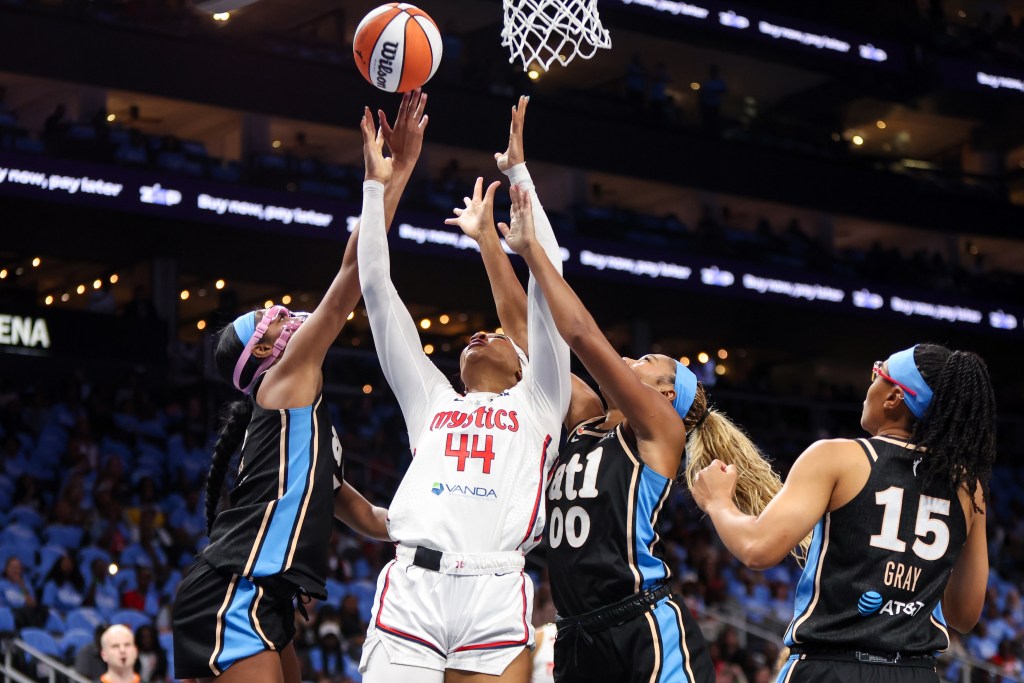 Washington Mystics forward Kiki Iriafen reaches for a rebound near the net with two hands. Two Atlanta Dream players are also going for the ball, but one is going up with only one hand, and the other is behind Iriafen.
