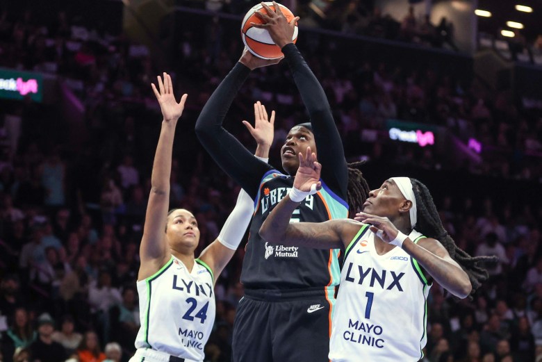 Jonquel Jones (center) jumps in her black Liberty jersey with the ball in her hands as Napheesa Collier (left) and Natasha Howard (right) defend beside her dressed in white