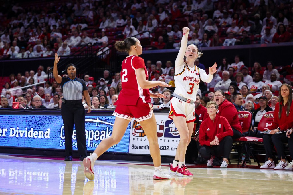 Addi Mack in a white shirt shoots the ball as a coach in red pullover kneels and watches and a player in a red shirt runs past.