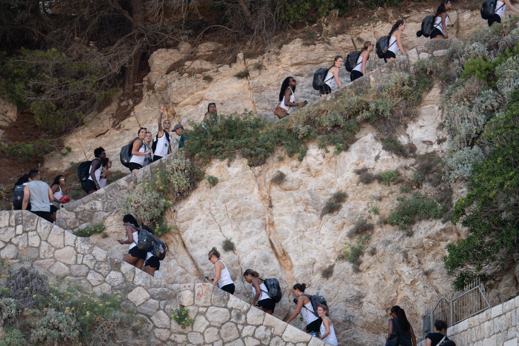 Members of the Harvard women's basketball program walk up steps in Dubrovnik, Croatia. Those toward the front of the line are walking up and to the right in the photo, and those toward the back of the line are walking up and to the left before reaching a turn. Many of them are carrying gray backpacks on their backs.
