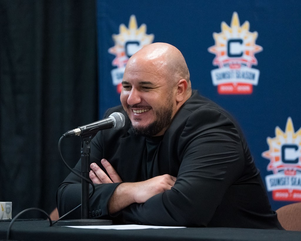 Rachid Meziane wearing black smiles with his arms crossed as he sits in front of a Connecticut Sun logoed backdrop and a microphone during his final press conference of the season.
