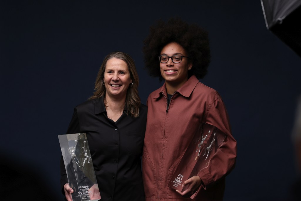 Cheryl Reeve (left) and Olivia Miles (right) wearing dark shirts in front of a dark backdrop holding large trophies and smiling wide