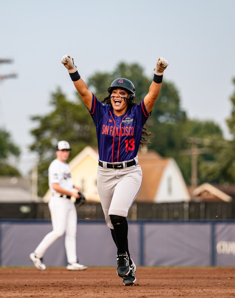 Firebells shortstop Joely Leguizamon is mid-step with her fists in the air and a big open-mouthed grin as she rounds the bases on her home run in Saturday's Game 3. Leguizamon is wearing a purple jersey with orange trim and "San Francisco" in orange across the front and her number 13 below the end of "Francisco." She also wears gray pants with purple trim cuffed to the knee, black socks and black spikes, black wrist bands, white batting gloves, and a black batting helmet with the Firebell's purple-and-orange logo. She has two streaks of eye black on each cheek a purple bruise (black eye) circles her left eye. Out of focus in the background, Queens shortstop Caitlin Eynon, in an all-white uniform, looks on.