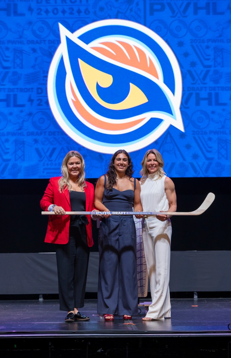 Three women stand holding a wooden hockey stick.