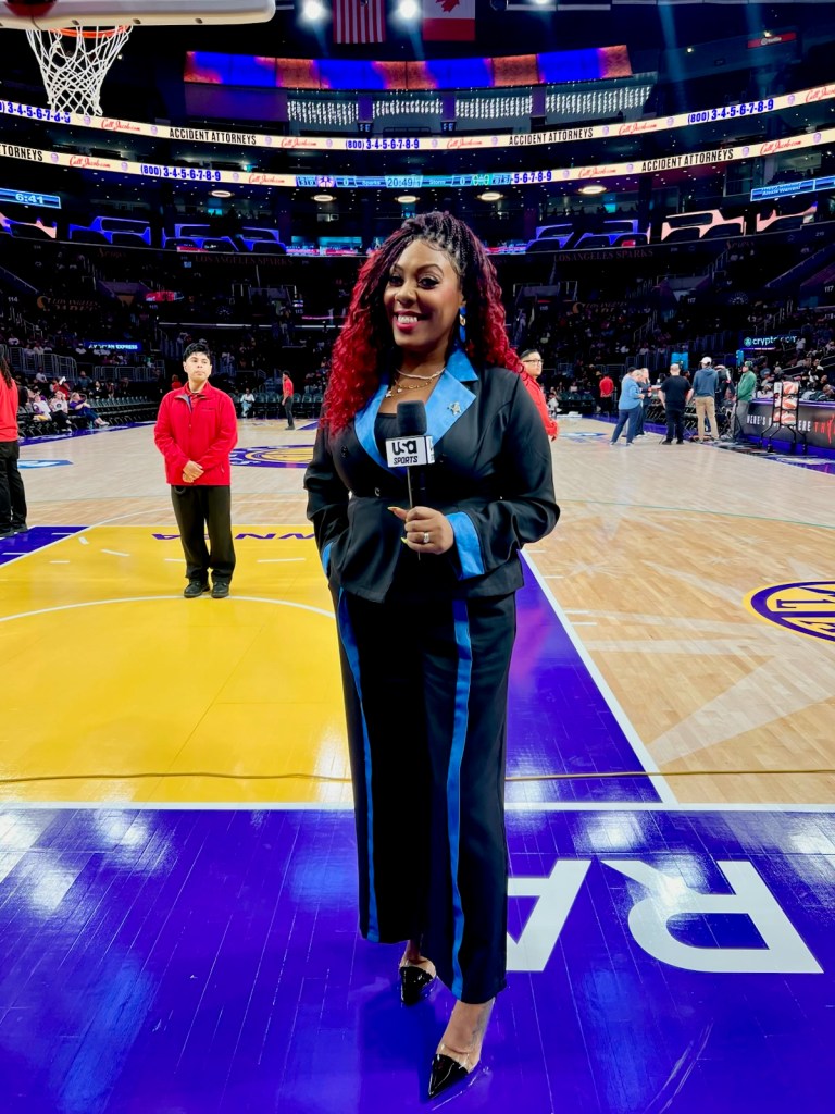 Terrika Foster-Brasby stands in a black outfit holding a microphone and smiling on a basketball court with the arena in the background.