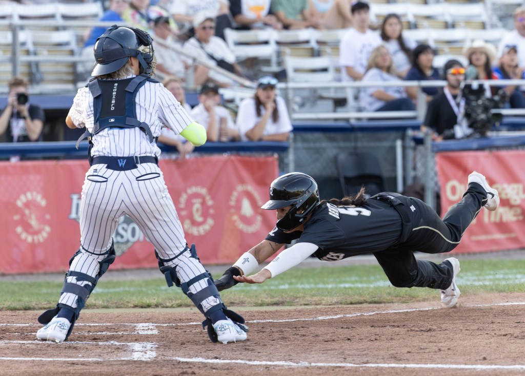 Heights catcher Denae Benites, seen from behind, awaits the throw at the plate while the Queens' Samaria Benítez dives, arms outstretched, toward home plate. Benites wears a pinstriped uniform and navy catcher's gear with a fluorescent yellow compression sleeve on her right arm and white spikes. She still has her catchers mask over her face. Benítez wears an all-black uniform with white and gold trim, a black batting helmet, white spikes, a white compression sleeve on her left arm, a white wristband on her right arm, and a black sliding mitt on her right hand.