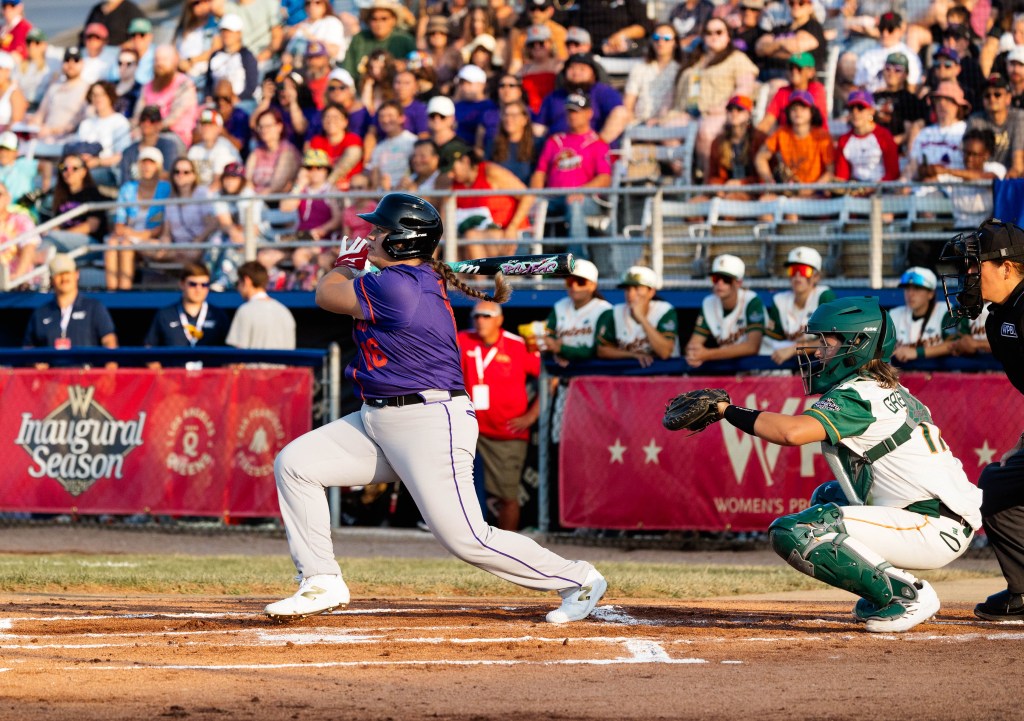 Andréanne Leblanc, batting lefty and seen from the third-base side of home plate, completes the followthrough of her swing while Heights catcher Beth Greenwood, in her squat, watches the flight of the ball to the right of the photo. Leblanc wears a purple jersey with orange trim, grey pants with purple trim, white spikes, a black batting ghelment, white and red batting gloves, and her braided brown pony tail whips out behind her back. Greenwood wears a white uniform with green sleeves and orange trim and green catcher's gear, including a hockey-style helmet. The umpire is at the edge of the photo the right. In the background, out of focus, the Hunters watch from their dugout behind red WPBL banners and the crowd watches above them.