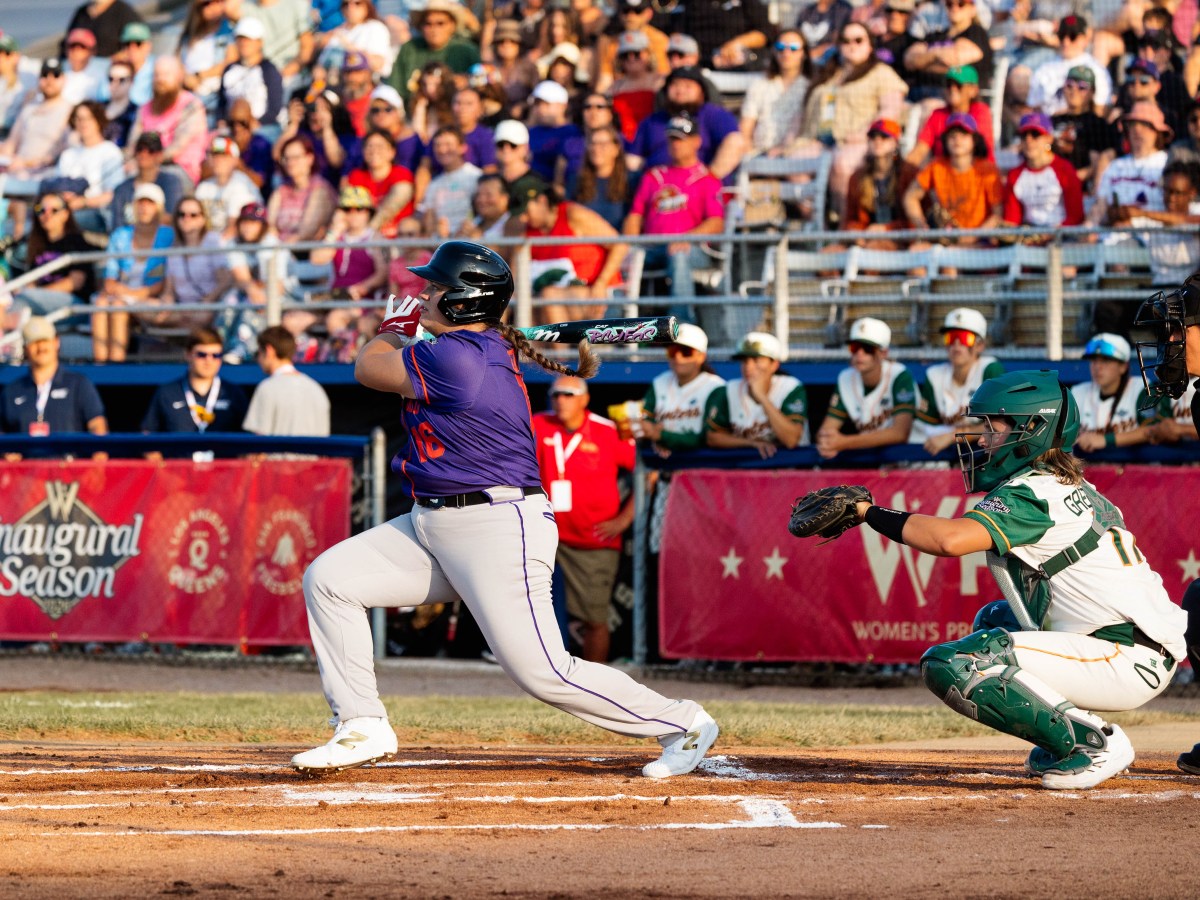 Andréanne Leblanc, batting lefty and seen from the third-base side of home plate, completes the followthrough of her swing while Heights catcher Beth Greenwood, in her squat, watches the flight of the ball to the right of the photo. Leblanc wears a purple jersey with orange trim, grey pants with purple trim, white spikes, a black batting ghelment, white and red batting gloves, and her braided brown pony tail whips out behind her back. Greenwood wears a white uniform with green sleeves and orange trim and green catcher's gear, including a hockey-style helmet. The umpire is at the edge of the photo the right. In the background, out of focus, the Hunters watch from their dugout behind red WPBL banners and the crowd watches above them.