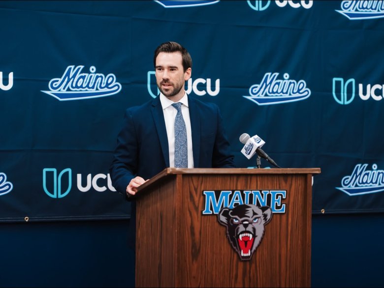 Alex Gettens speaks behind the podium at Maine in front of a Maine branded backdrop