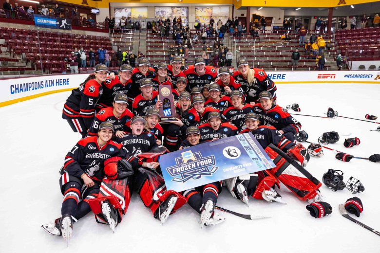 Northeastern’s women’s hockey team celebrates on the ice in a group photo with a regional final trophy and Frozen Four ticket.