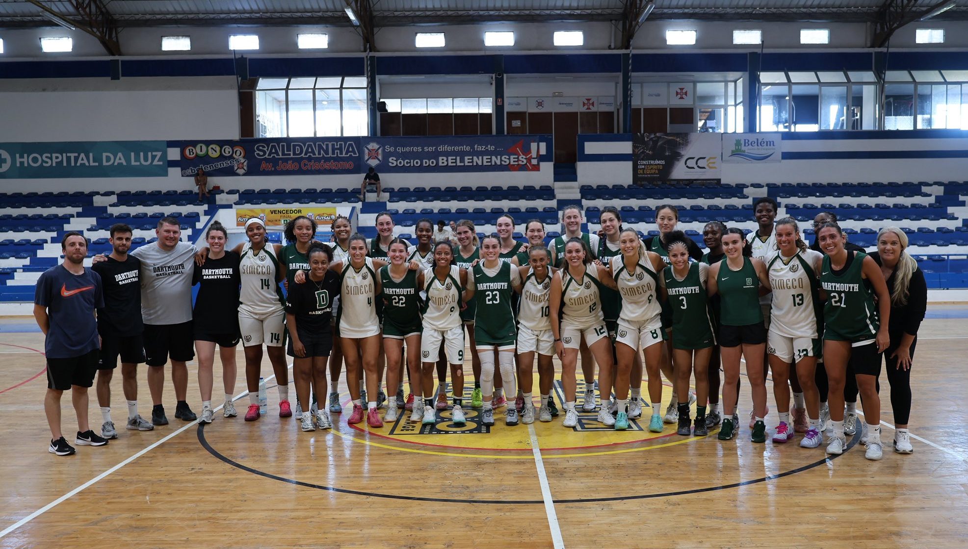 Dartmouth players and coaches stand at center court with players from Simecq, a Portugese club team. They smile for a photo, and many of those pictured have their arms around one another. The Dartmouth players are wearing green jerseys, and the Simecq players are in white jerseys.
