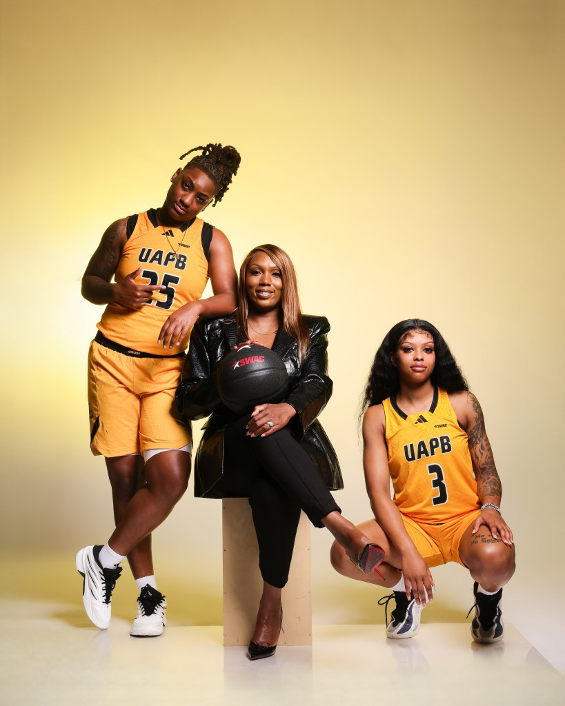A player in a gold jersey is standing with her arm on a coaches shoulder in a black jacket who is sitting with her leg crossed and another player in a gold jersey kneeling.