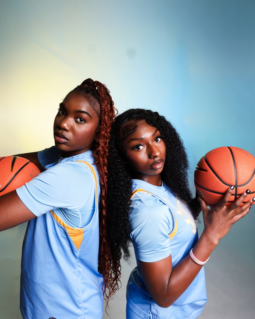 Two basketball players in Columbia Blue uniforms stand back to back with the one on the left cradling a ball and the the one on the right holding the ball.