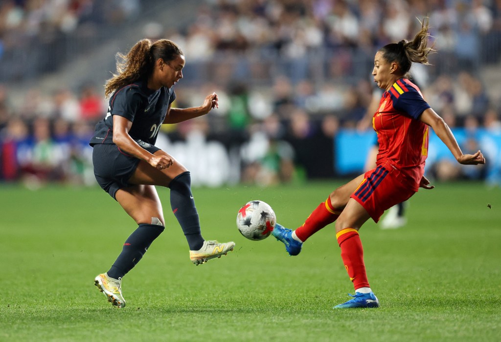 A Spain player in a red uniform kicks a ball while a U.S. player in a blue shirt goes after it.