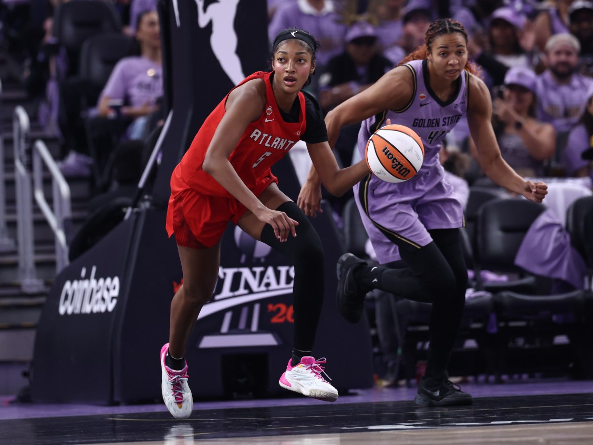 Two players battle for the basketball on a court, one in a red jersey and one in a purple jersey.