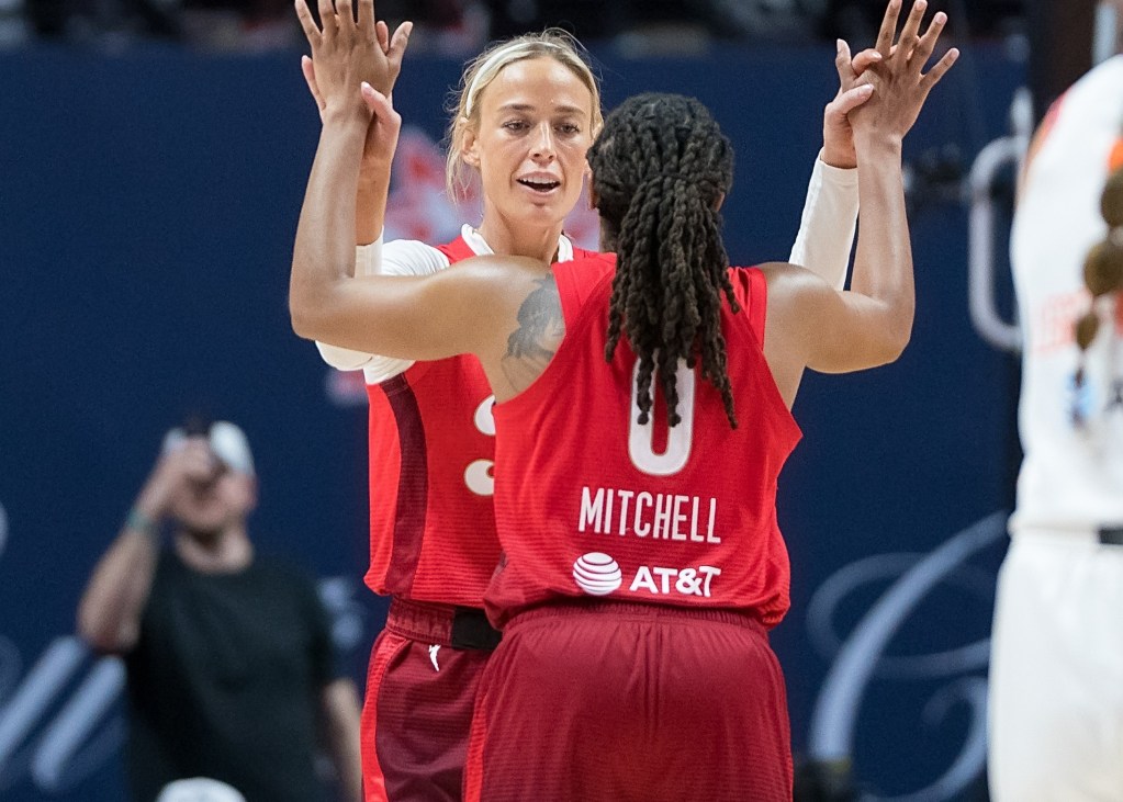 Indiana Fever players Kelsey Mitchell and Sophie Cunningham high-five with both hands during a game. Cunningham is facing the camera, while Mitchell is facing Cunningham with her back to the camera.
