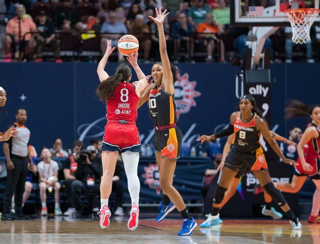 Washington Mystics point guard Georgia Amoore is shown with her back to the camera, shooting a right-handed jump shot. Connecticut Sun center Olivia Nelson-Ododa closes out on Amoore, extending her left arm up to try to block the shot or limit Amoore's vision.