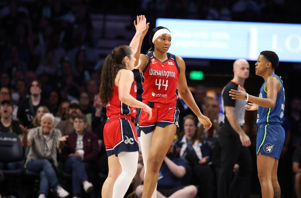 Washington Mystics point guard Georgia Amoore is shown from the side. She reaches her left arm up to high-five forward Kiki Iriafen, who is facing the camera. In the background, Minnesota Lynx guard Courtney Williams holds her hands out in confusion.