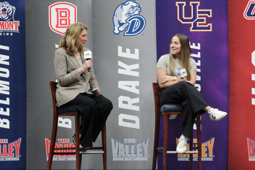 A basketball analyst interviews a player on site at a media day. They are both sitting in high-boy chairs. 