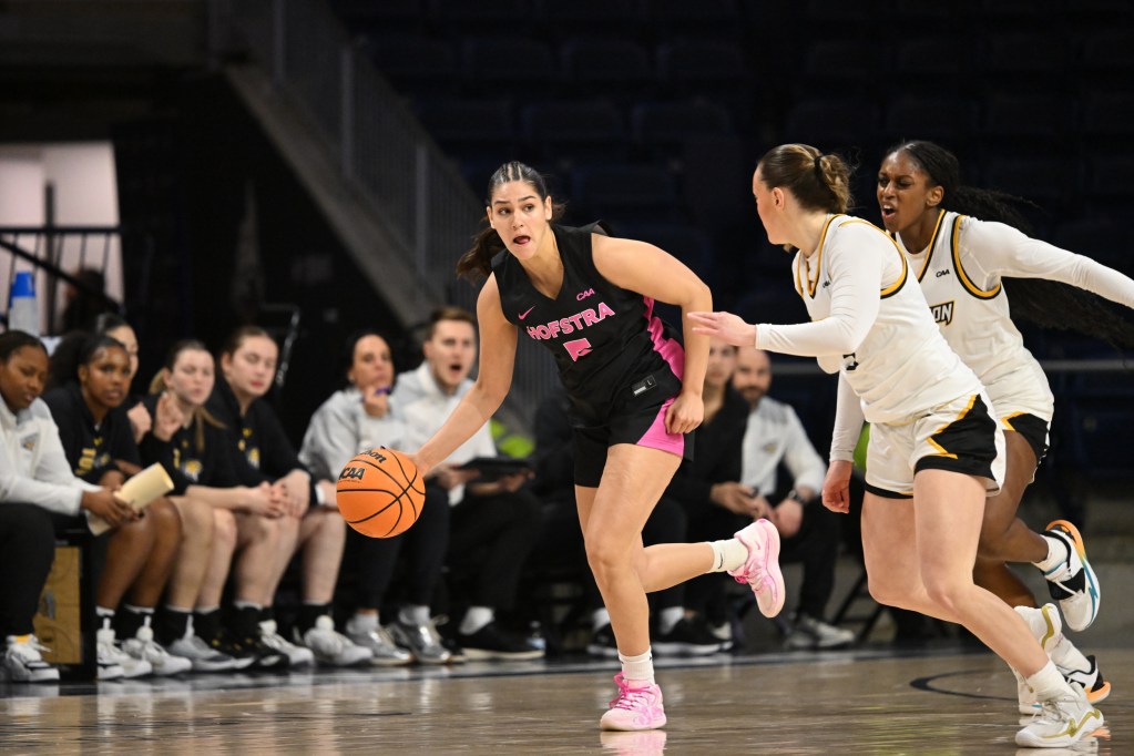 A basketball player in a black uniform dribbles despute pressure from two players in white uniforms.