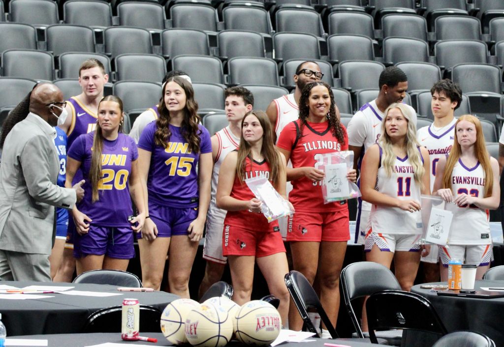 Male and female basketball players in purple, red and white jerseys gather in the stands of an arena. 