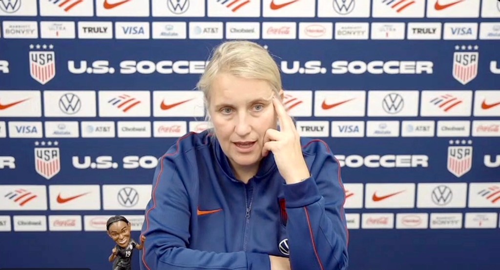 United States women's national team head coach Emma Hayes sits in front of a U.S. soccer backdrop and addresses the media over Zoom. She rests her left hand on the side of her face as she talks, and her right arm rests on the table in front of her. Near her right elbow, a bobblehead of retired player Crystal Dunn is visible.