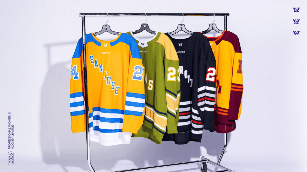 The four new PWHL jerseys hang on a rack. San Jose, i orange and blue is first, followed by Las Vegas in green and ochre, Detroit in blue, red and white, and Hamilton with burgundy and mustard.